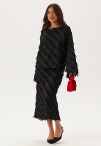Svart tvådelad outfit bestående av en långärmad topp och en knälång kjol, båda prydda med vertikala fransdetaljer. Accentuerad med en röd clutch.