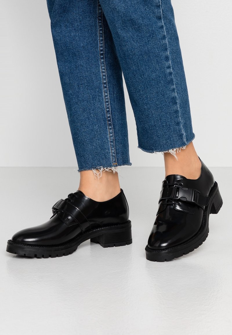 Jambes portant un jean bleu coupé et des chaussures noires en cuir brillant avec des talons épais et des brides à boucle sur un sol lisse et clair.