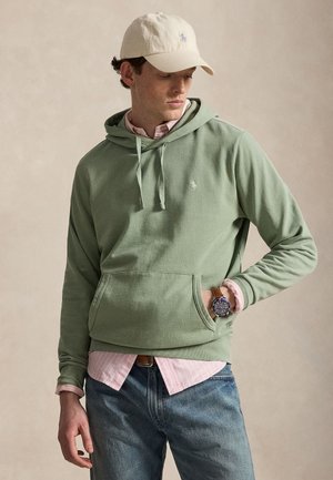 Mand iført en lysegrøn hættetrøje over en lyserød skjorte, blå jeans, beige kasket og armbåndsur, stående med den ene hånd i hættetrøjlommen.