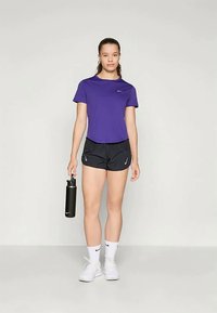 Atletska ženska stoji s črno steklenico za vodo, oblečena v vijolično majico, črne kratke hlače, bele nogavice in bele superge pred enobarvno ozadjem.