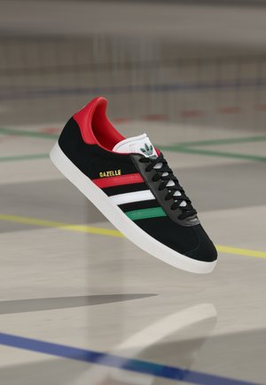 Czarne zamszowe sneakersy z czerwonymi akcentami, białymi i zielonymi paskami oraz czarnymi sznurowadłami. Posiadają białą podeszwę i złoty napis "GAZELLE" po boku.