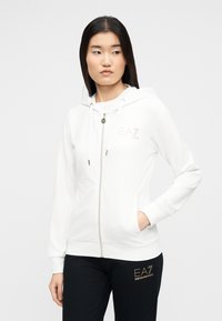 Νεαρή γυναίκα που φοράει λευκό φούτερ με φερμουάρ και κουκούλα και μαύρο παντελόνι, στέκεται με το ένα χέρι στην τσέπη, κοιτάζοντας πλάγια.