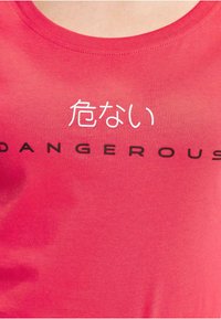 Red cotton t-shirt with a round neckline. Features black "DANGEROUS" text and white Japanese characters. Smooth texture with a fitted shape.