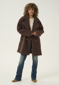 Pruun tekstureeritud villane mantel laia krae, topeltääkli disain ja suured nööbid. Kombineeritud siniste laiade teksade ja beežite saapadega.