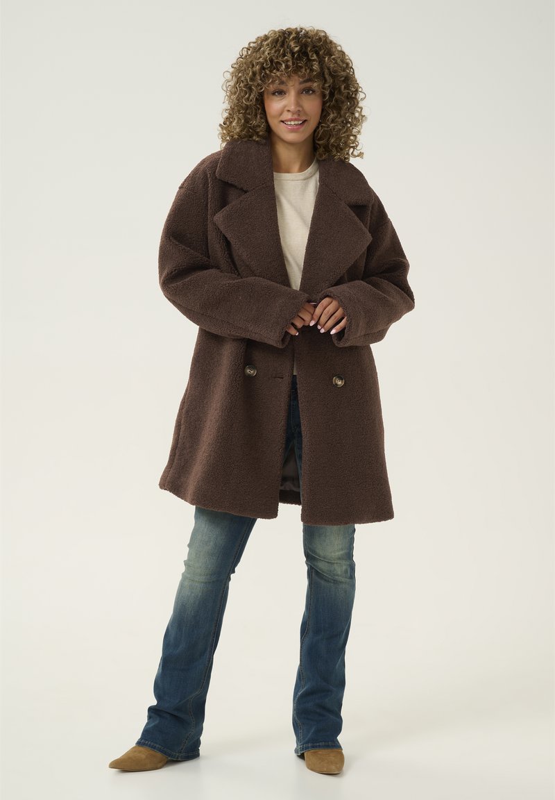 Brauner, strukturierter Wollmantel mit breitem Revers, doppelt knöpfbarem Design und großen Knöpfen. Kombiniert mit blauen, ausgestellten Jeans und beigen Stiefeln.