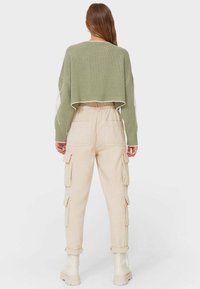 Pull en mailles vertes à manches longues, associé à un pantalon cargo beige avec poches latérales et bas de pantalon retroussés. Les chaussures sont des bottes robustes.