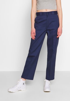 Persoon die marineblauwe, recht gesneden broek draagt, witte hoge sneakers en een grijs mouwloos cropped topje, staand tegen een effen witte achtergrond.