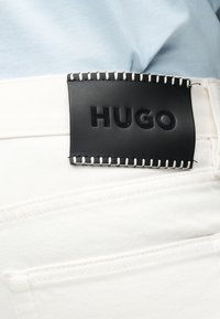 Μαύρη ραμμένη δερμάτινη ετικέτα με εντυπωμένο το "HUGO" σε λευκό τζιν τζιν φόρεμα, που φοριέται με ανοιχτό μπλε τοπ.