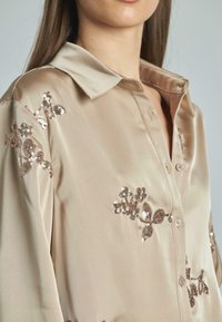 Camicetta beige in satin con colletto, caratterizzata da ricami floreali e paillettes marroni sulle spalle e sul petto, chiusura con cinque bottoni.