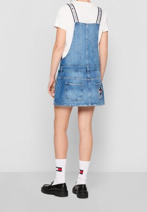 Person iført en blå denim overallkjole med mærkede stropper, hvid T-shirt, hvide sokker med logo og sorte loafers, stående med ryggen mod.