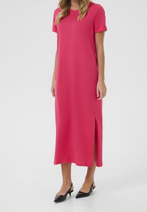 Frau trägt ein knallpinkes Midikleid mit kurzen Ärmeln und seitlichem Schlitz, kombiniert mit schwarzen spitzen Slingback-Pumps, stehend vor weißem Hintergrund.