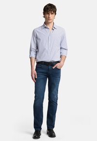 Camicia button-up con righe blu e bianche, abbinata a jeans blu scuro e scarpe nere. Il modello sta in piedi con le mani in tasca contro uno sfondo bianco.