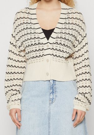 Femme portant un cardigan en maille rayé crème et noir avec des boutons, sur un haut noir et une jupe en jean bleu clair.