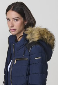 Giacca imbottita blu navy con bordo in pelliccia sintetica sul cappuccio, dettagli in pelle nera sulle spalle, tasche con zip e dettagli in hardware dorato.