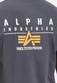 Hallhallane puuvillane T-särk, millel on suur valge ja kollane graafika, kus on tekst "ALPHA INDUSTRIES", ja seljal tiibadega embleem.