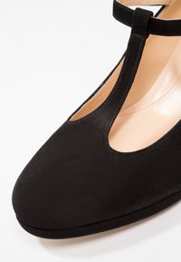 Zapato de ante negro con punta redonda y un diseño de tira en T. Presenta un detalle troquelado cerca de la punta y un forro interior suave.