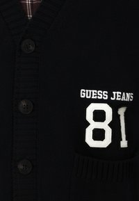 Jūrinis megztinis su sagų uždarymu, su iškilnios tekstūros raštu, išsiuvinėtu „GUESS JEANS“ ir „81“ baltos spalvos, su kišene priekyje.