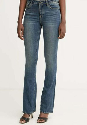 Donna che indossa jeans blu a vita alta e gambe dritte e sandali neri aperti con tacco, in piedi davanti a uno sfondo semplice.