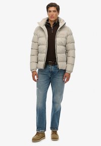 Chaqueta acolchada gris claro con capucha, cierre delantero, puños elásticos y dos bolsillos laterales. Lleva puesta sobre un suéter marrón oscuro y jeans azules.