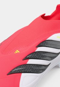 Κόκκινο ποδοσφαιρικό παπούτσι με εφαρμοστό πάνω μέρος, υφαντή επιφάνεια, μαύρες και λευκές λεπτομέρειες και σχέδιο στο πλάι. Διακοσμημένο με κίτρινο λογότυπο.