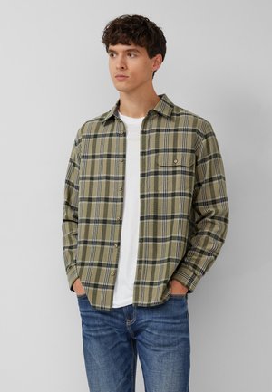 Jeune homme aux cheveux bruns courts portant une chemise à carreaux vert olive boutonnée par-dessus un t-shirt blanc et un jean bleu, les mains dans les poches.