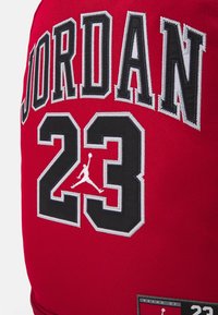 Czerwony materiał z czarnym i białym haftowanym napisem "JORDAN" nad dużą liczbą "23" z logo Jumpman wewnątrz "3" na plecaku.