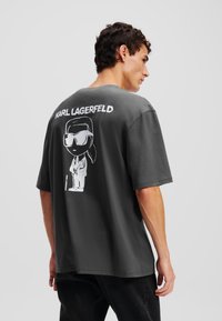 Sivá oversized tričko s krátkymi rukávmi, na zadnej strane s bielou kreslenou grafikou a textom "KARL LAGERFELD". Hladká bavlnená textúra.