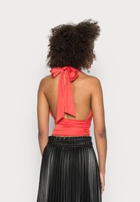 Top vermelho com alças de halter e um laço largo nas costas, detalhado com franzidos, combinado com uma saia preta plissada. Textura de tecido suave.