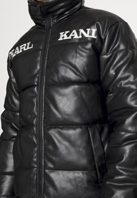 Czarna kurtka puchowa o błyszczącej fakturze, z pikowanymi sekcjami, wysokim kołnierzem oraz haftowanymi logotypami "KARL" i "KANI" z przodu.