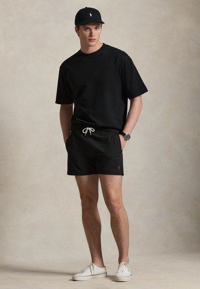 Νεαρός άνδρας που φοράει μαύρο καπέλο, μαύρο φαρδύ κοντομάνικο, μαύρα σορτς με λευκό κορδόνι, ρολόι και λευκά αθλητικά παπούτσια, στέκεται μπροστά σε απλό φόντο.