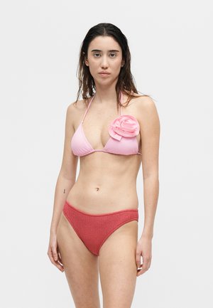 Γυναίκα με μακριά σκούρα μαλλιά φορά ροζ μπικίνι τοπ με μεγάλο λουλουδάτο διακοσμητικό και κόκκινο υφασμάτινο κάτω μέρος μπικίνι, στέκεται απέναντι σε απλό φόντο.