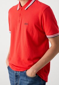Mężczyzna w czerwonej koszulce polo BOSS z białymi i czarnymi paskami przy kołnierzyku i rękawach, z rękami w kieszeniach niebieskich dżinsów, widoczna jest jego klatka piersiowa.
