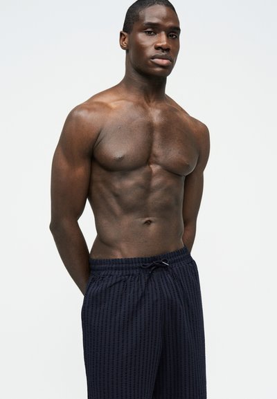 Hombre musculoso sin camisa de piel oscura, vestido con pantalones cortos azul marino con cordón, de pie con las manos detrás de la espalda contra un fondo liso.