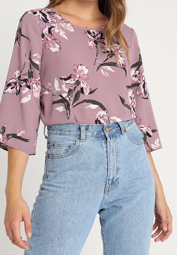 Femme portant un chemisier mauve à fleurs avec des manches trois-quarts, rentrée dans un jean taille haute bleu clair.