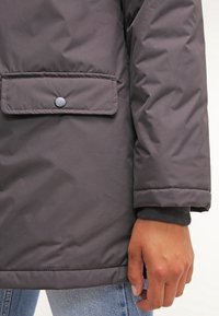 Gray, padded jacket with a snap-button pocket, ribbed cuffs, and smooth, water-resistant material. Close-up of the sleeve and pocket.