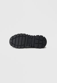 Schwarze Gummisohle mit tiefen Profilmustern und Rillen, ausgestattet mit einer abgerundeten Zehenpartie und einem mittig erhabenen Logo auf der Zwischensohle.