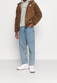 Bruine corduroy zip-up jas over een grijze hoodie, gecombineerd met losse lichtblauwe jeans en witte sneakers met gouden accenten.
