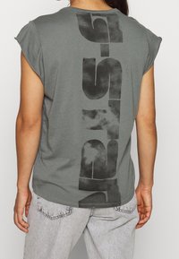 Persona vistiendo una camiseta sin mangas gris con un gran estampado vertical negro y jeans gris claro, mostrada desde atrás.