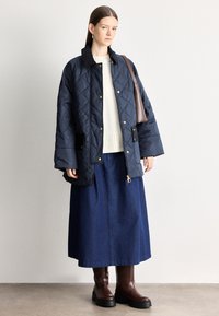 Marineblauwe doorgestikte jas met gouden knopen, gecombineerd met een crème gebreide trui en een blauwe midi-rok. Bruine laarzen met grove hak maken de outfit compleet.