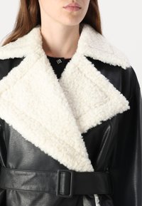Sort læderjakke med hvid faux shearling krave, bæltedesign og tekstureret finish. Bemærkelsesværdig trekantet kraveform og spænde-detail.