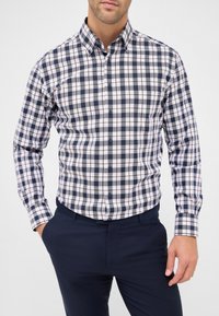 Man in een knopenblauw, wit en bruin geruit overhemd met lange mouwen en blauwe broek, met de rechterhand in de zak, tegen een effen lichte achtergrond.