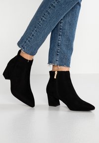 Sorte ruskinds ankelstøvler med spids tå, blokhæl og lynlås på siden, prydet med guldfarvet hardware. Båret med blå jeans med frynset kant.