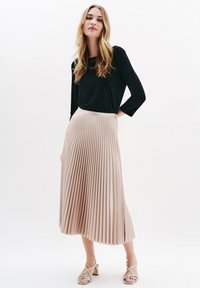 Fältete Midi-Rock in hellbeige mit eng anliegendem Bund, kombiniert mit einem schwarzen Langarmtop. Zeigt eine weiche Textur und Bewegung.