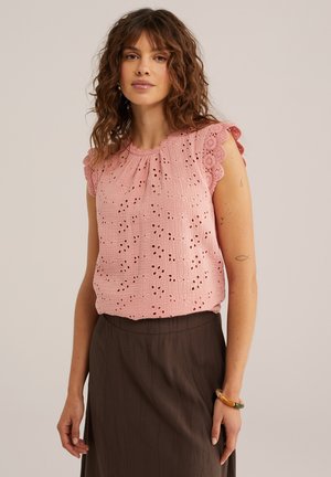 Mujer con cabello rizado que lleva una blusa rosa sin mangas con ojales y mangas de encaje con bordes ondulados, y una falda oscura de talle alto.