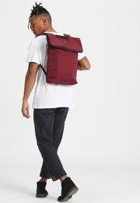 Mochila marrón con una tapa plegable y detalles en negro, hecha de tela resistente. Correas negras ajustables y un respaldo acolchado para mayor comodidad.