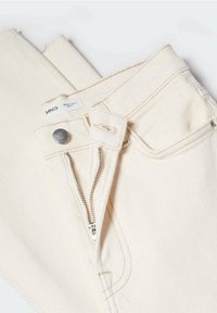 Jean en denim couleur crème avec une fermeture éclair ouverte, un bouton en métal et une étiquette de marque visible sur la ceinture.