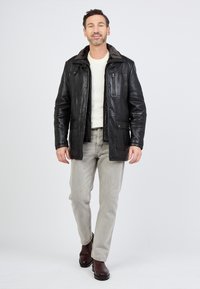 Zwart leren jack met een bontgevoerde kraag, knoop- en ritssluiting, meerdere zakken, gedragen over een wit shirt en grijze broek. Bruine laarzen.