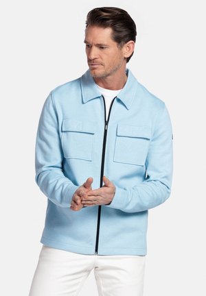 Homme aux cheveux bruns courts portant une veste zippée bleu clair avec deux poches poitrine et un pantalon blanc, les mains jointes.