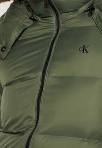 Oliwkowa pikowana kurtka z zamkiem błyskawicznym, kapturem i logo na piersi; posiada czarny zamek i błyszczącą, gładką fakturę.