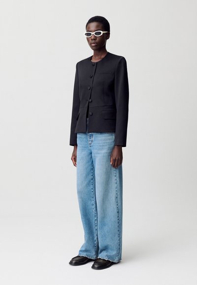 Una persona está de pie contra un fondo neutro, vistiendo una chaqueta negra a medida, unos vaqueros azules de gran tamaño y gafas de sol blancas.